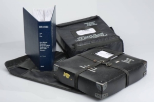 blue braille book standing upright on its black mailing case with two additional braille books in mailing cases lying nearby